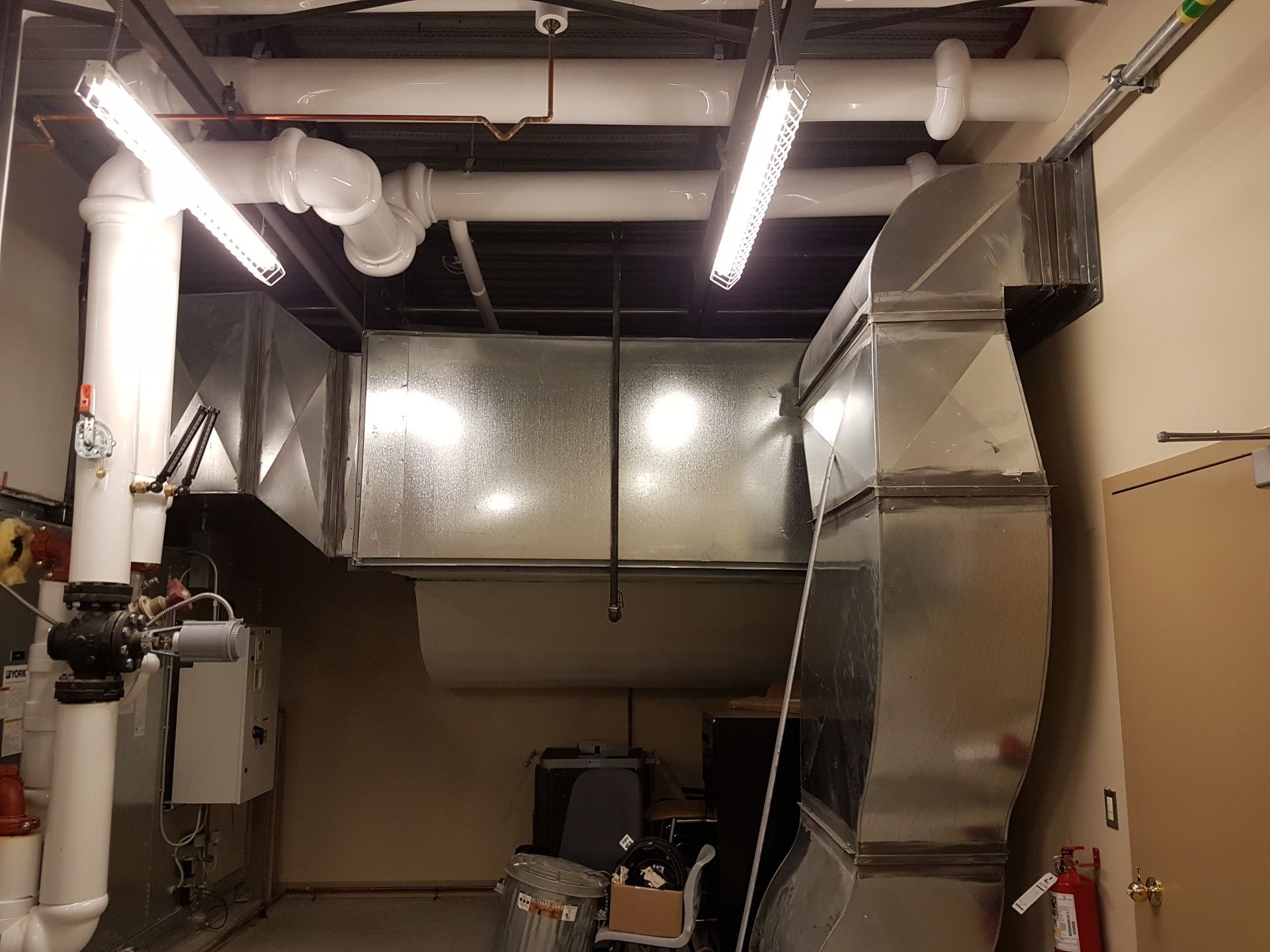 HVAC ductwork and pipes in a utility room, with lighting and a door on the right.