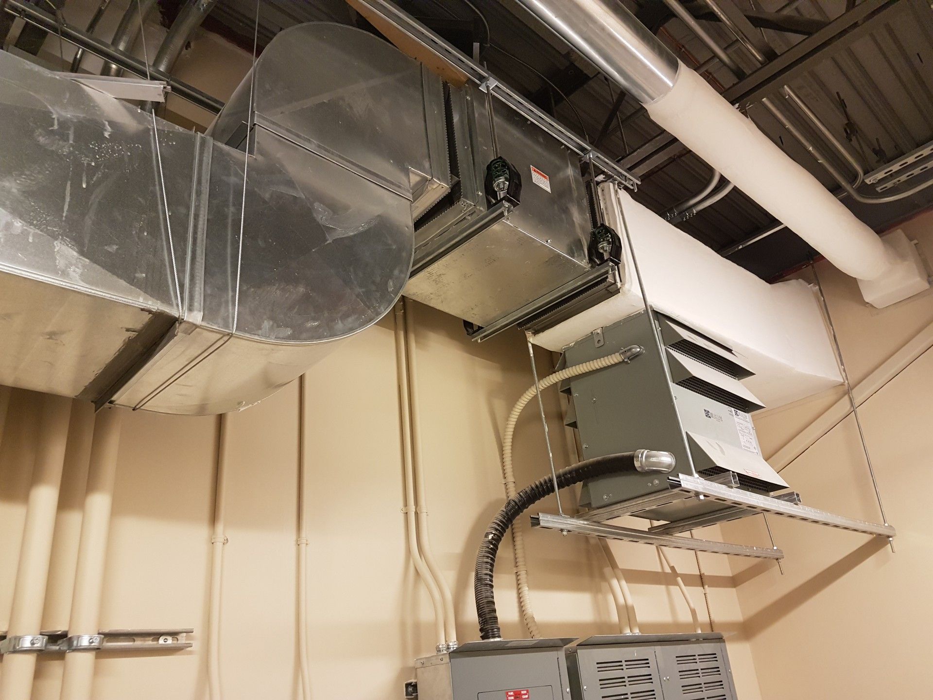 HVAC unit and ductwork hanging from a ceiling in a beige-walled room. Silver metal, white pipes, and wires are visible.
