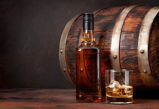 A whiskey bottle and a glass with ice cubes sit on a wooden surface in front of a rustic wooden barrel.