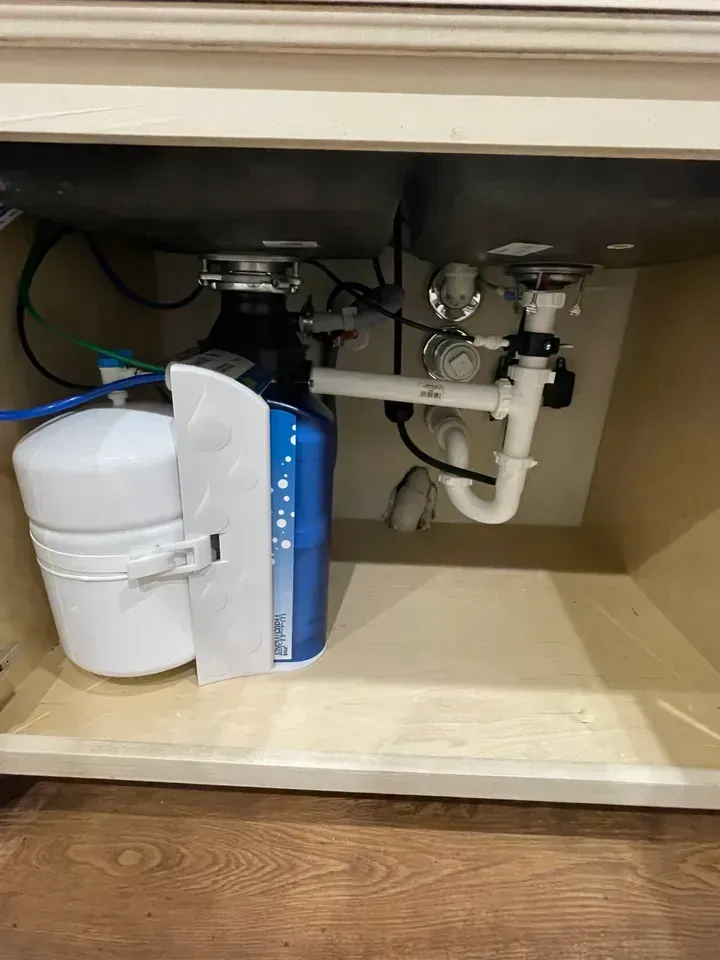 Under-sink view: Blue water filter, disposal, and plumbing, beige cabinet interior.