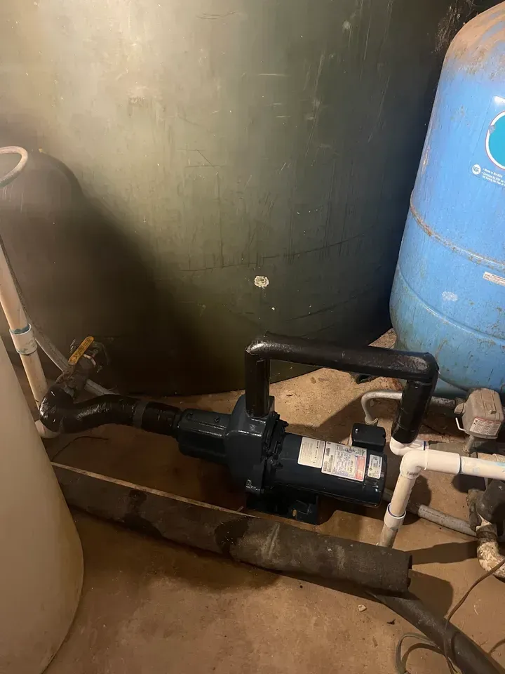 Dark blue water pump with black handle, connected to plumbing near tanks in a basement.