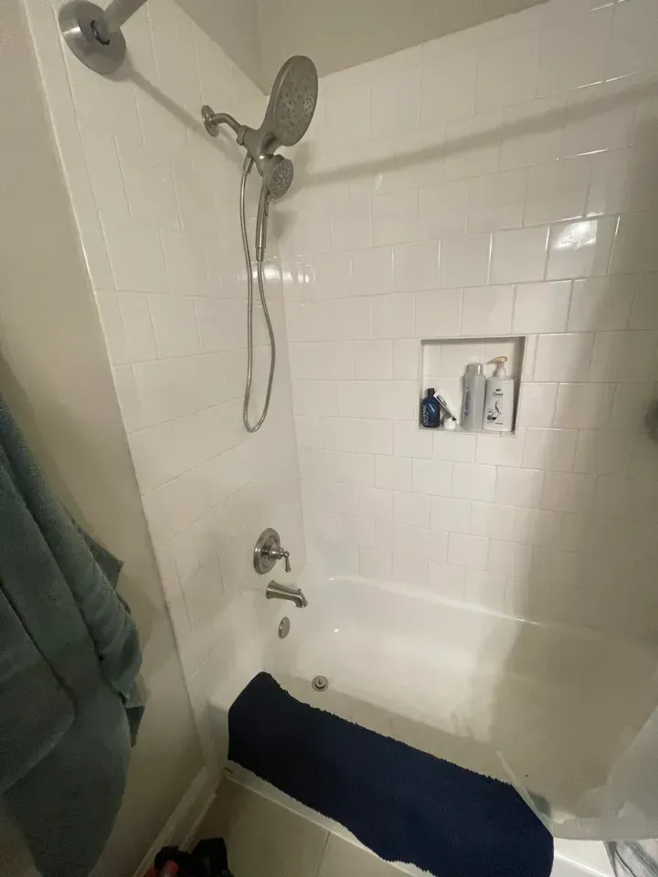 White tiled shower with multiple shower heads and a built-in shelf holding toiletries. A dark blue bath mat sits in the tub.