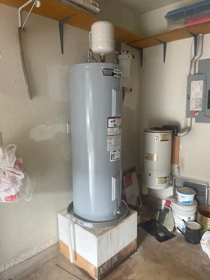 A tall, gray water heater on a concrete block in a garage, with other equipment and shelves.