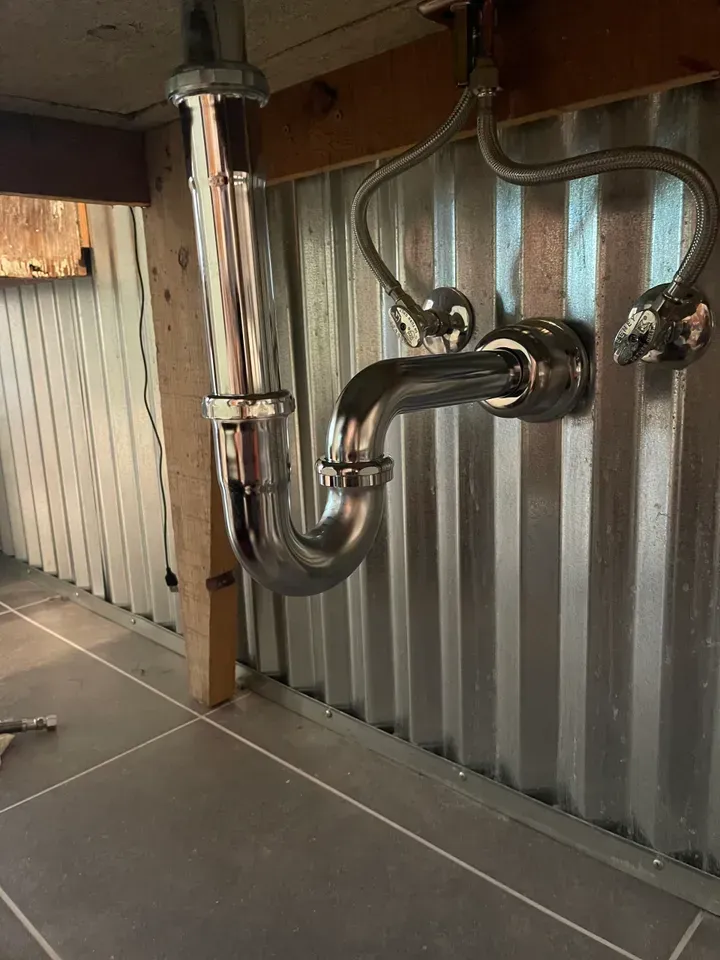 Chrome plumbing under a sink against corrugated metal wall, including P-trap and water supply lines.