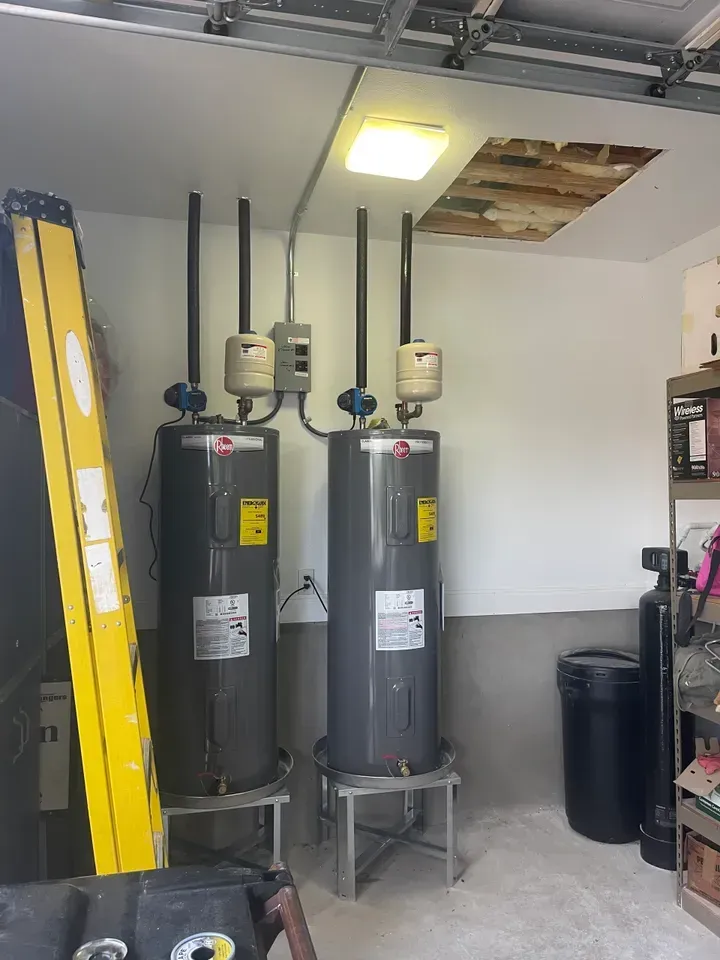 Two water heaters in a garage, with metal stands, pipes, and electrical box, with a damaged ceiling tile.