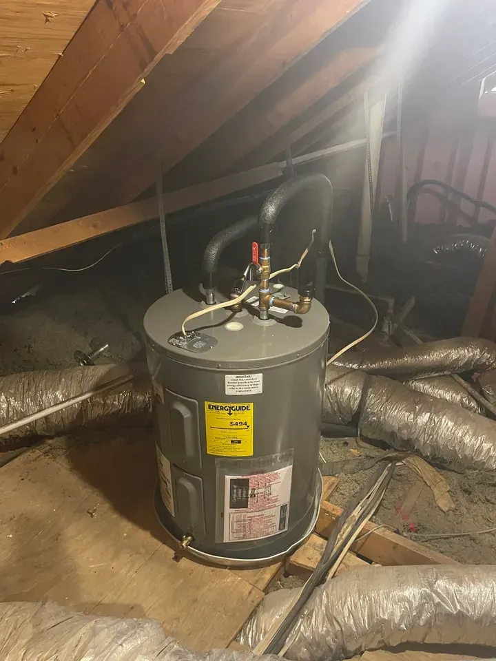 Water heater in an attic setting; gray tank with black pipes and yellow energy label, surrounded by insulation and wood beams.
