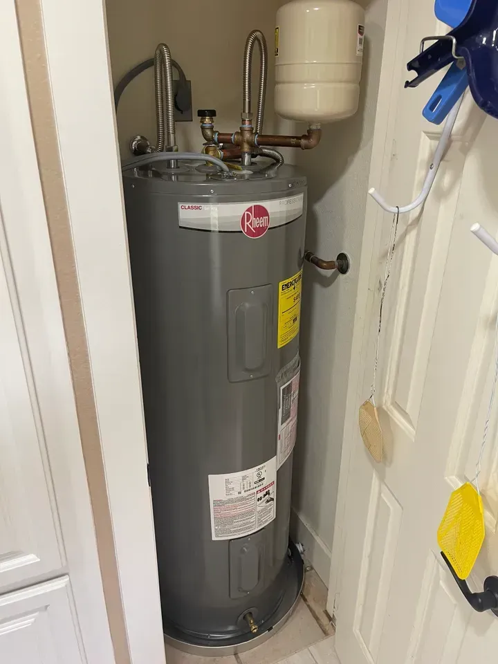 A tall, gray Rheem water heater in a small utility closet next to white shelving and hooks.