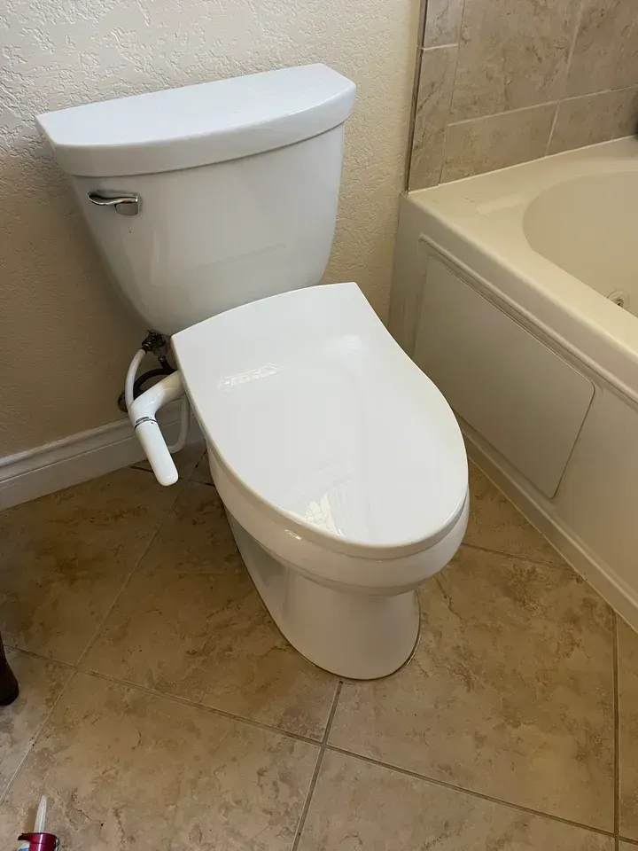 White toilet with a bidet attached, near a white bathtub, on tile floor.