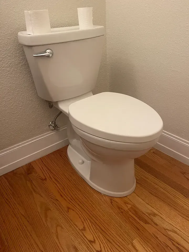 White toilet in a bathroom with wood flooring, rolls of toilet paper on tank, and white walls.
