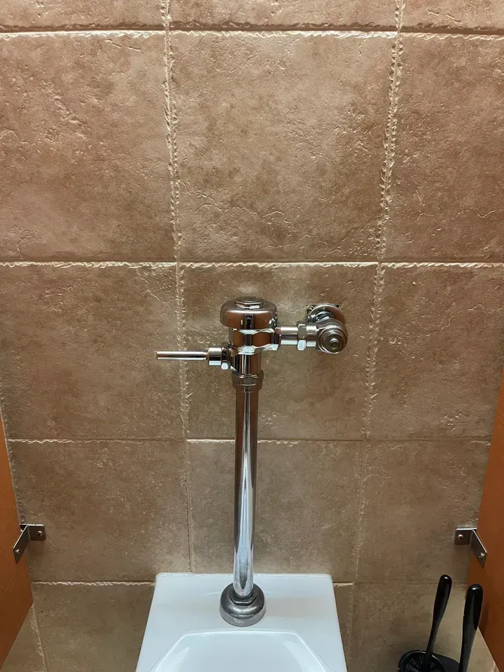 Toilet with a chrome flushometer against a beige tile wall.