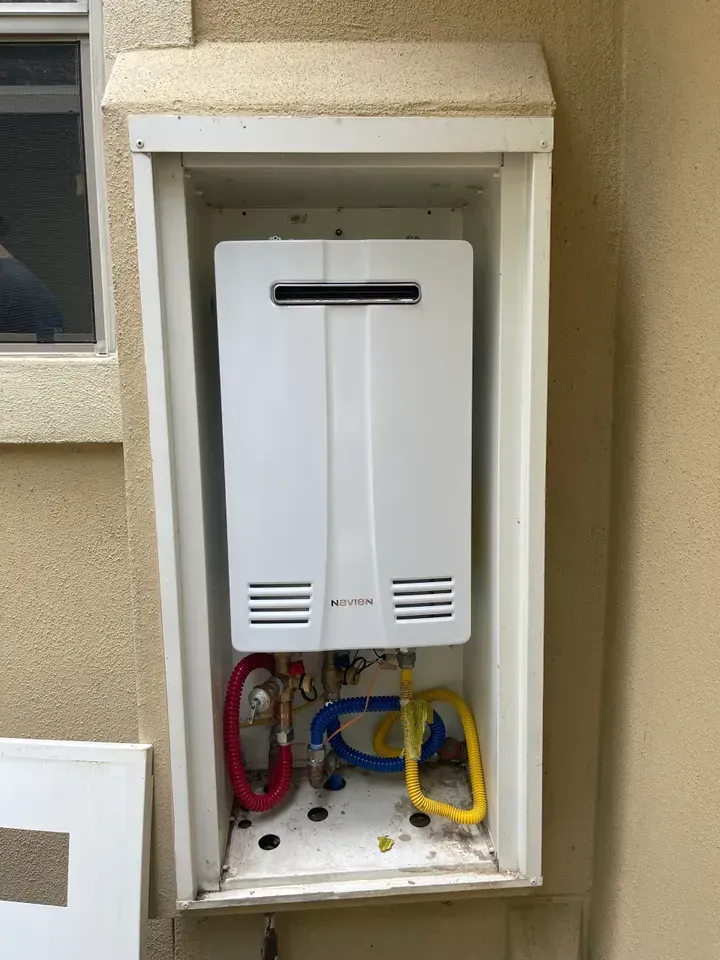 White tankless water heater inside a white metal box on an exterior wall; red and yellow gas lines visible.