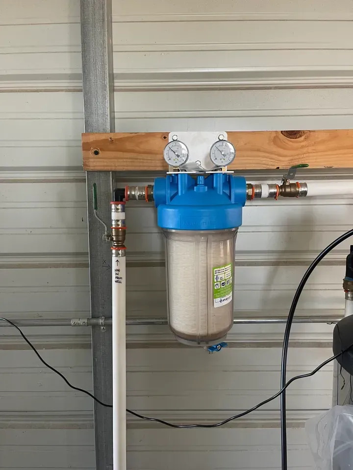 Water filter system mounted on a wooden board with pressure gauges and PVC pipes in a garage.