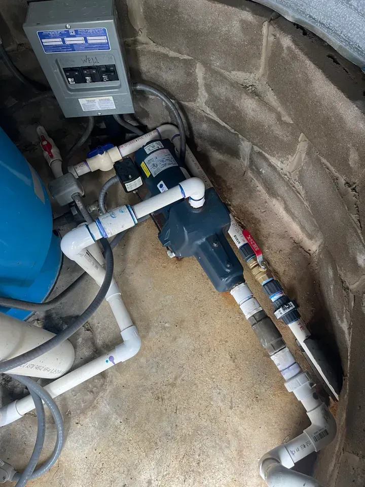 Water pump system in a stone-lined well. Electrical panel, blue tank, and white PVC pipes are visible.