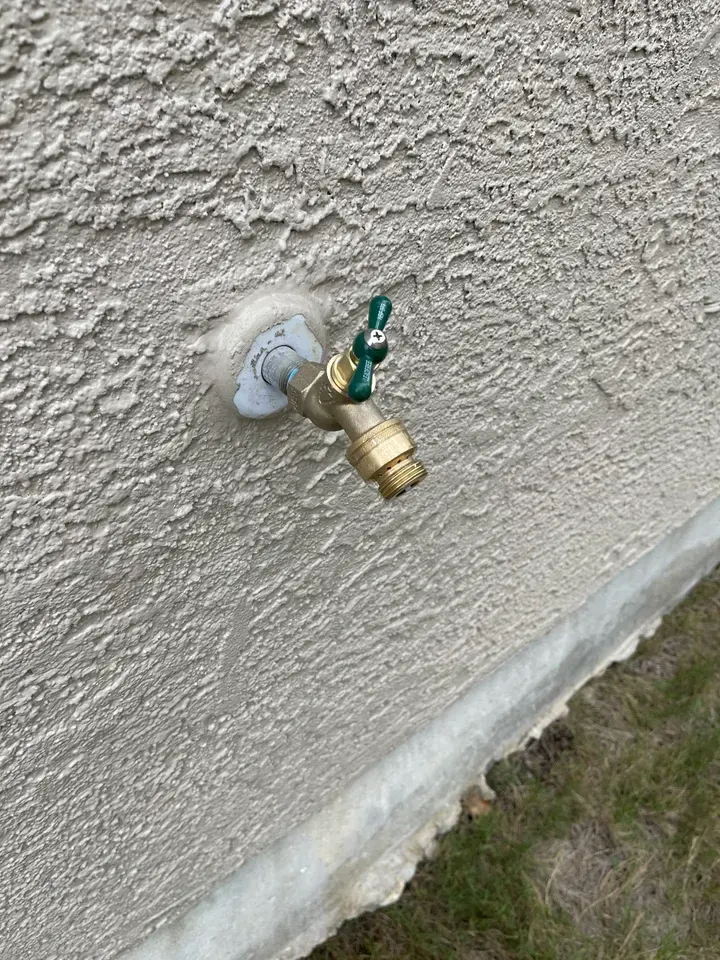 Outdoor water faucet on a textured light-colored wall with a green handle, angled downward.