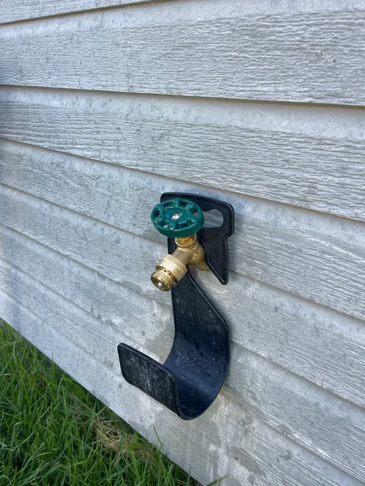 A green-handled water spigot with hose holder on a gray siding wall.