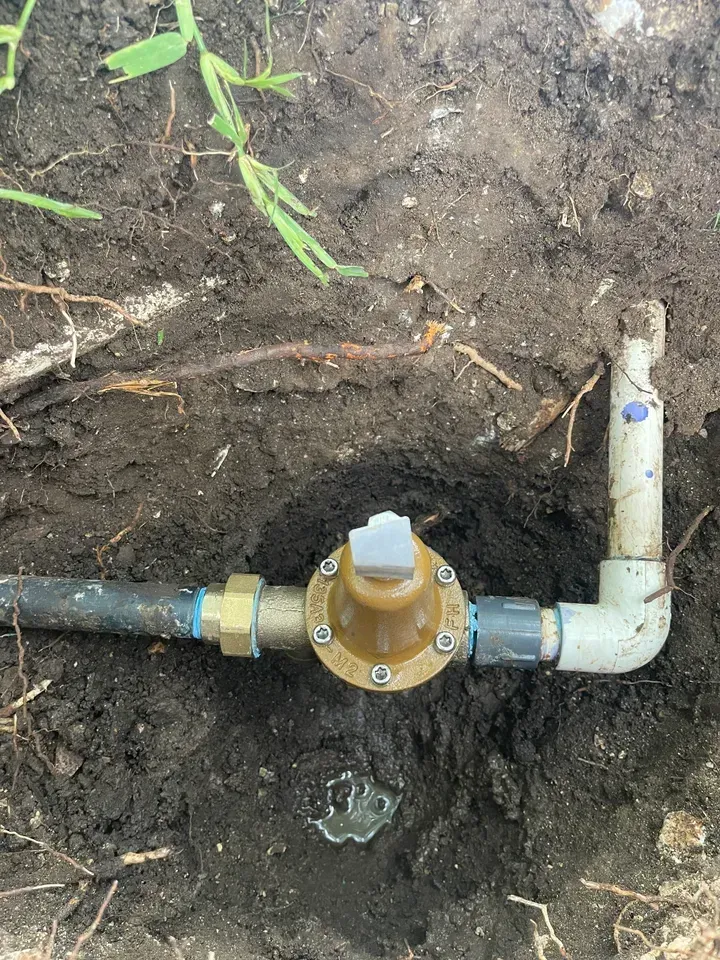 Sprinkler system valve in a dirt hole; brass and PVC pipes connect, brown dirt surrounds.