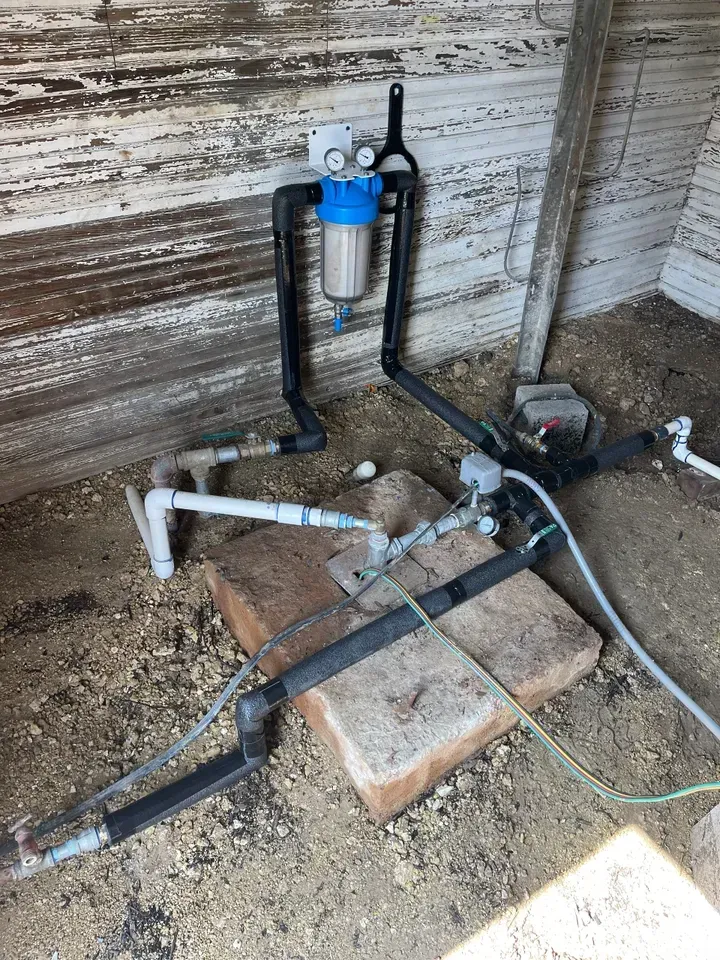 Water filtration system mounted on a wall. Black pipes, blue filter, and gauges, outside a weathered building.