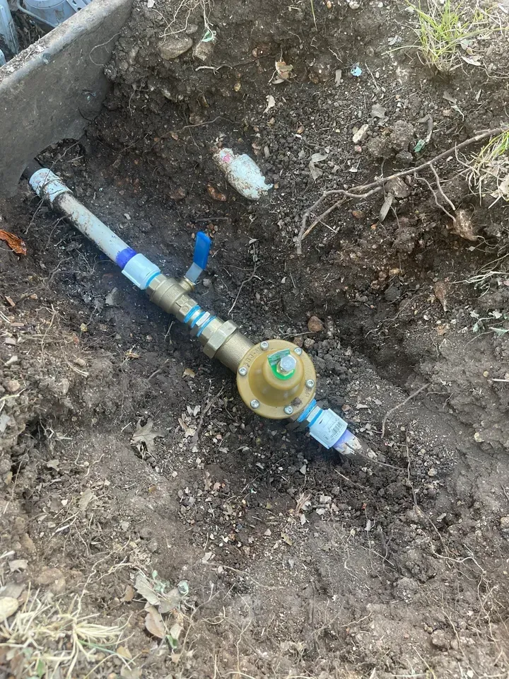 Water pressure regulator installed in dirt, with blue handle and metal piping.