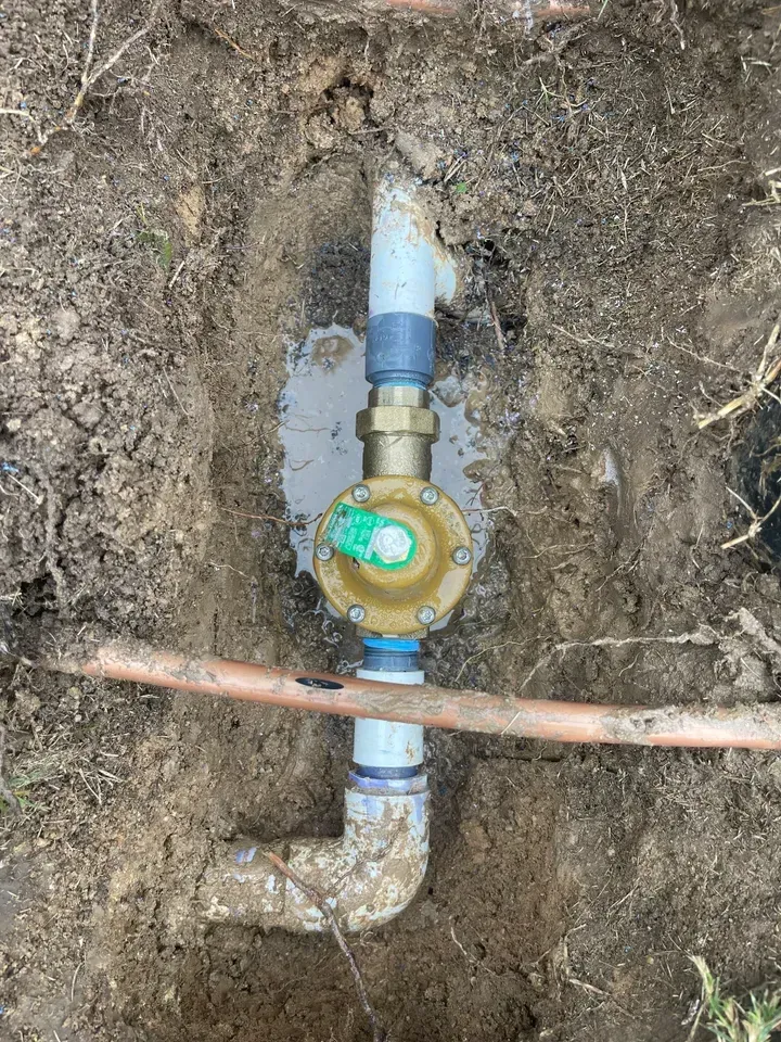 Sprinkler system valve in a dirt trench; valve is gold and green, with white and copper piping.