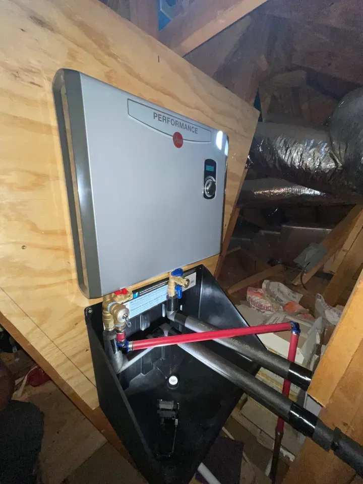 Gray water heater mounted on plywood in an attic, with plumbing and ductwork visible.