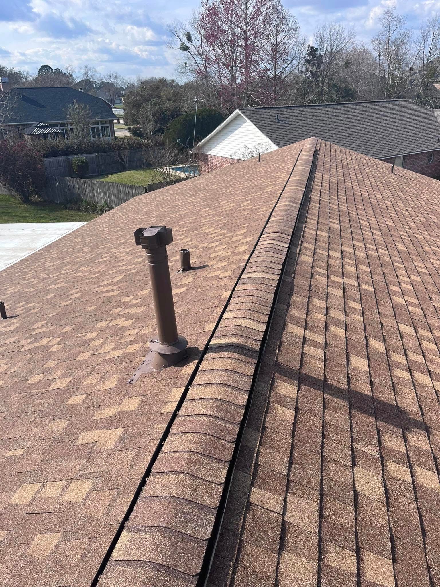 A brown shingle roof with a vent pipe and ridge cap, with surrounding houses and trees in view.