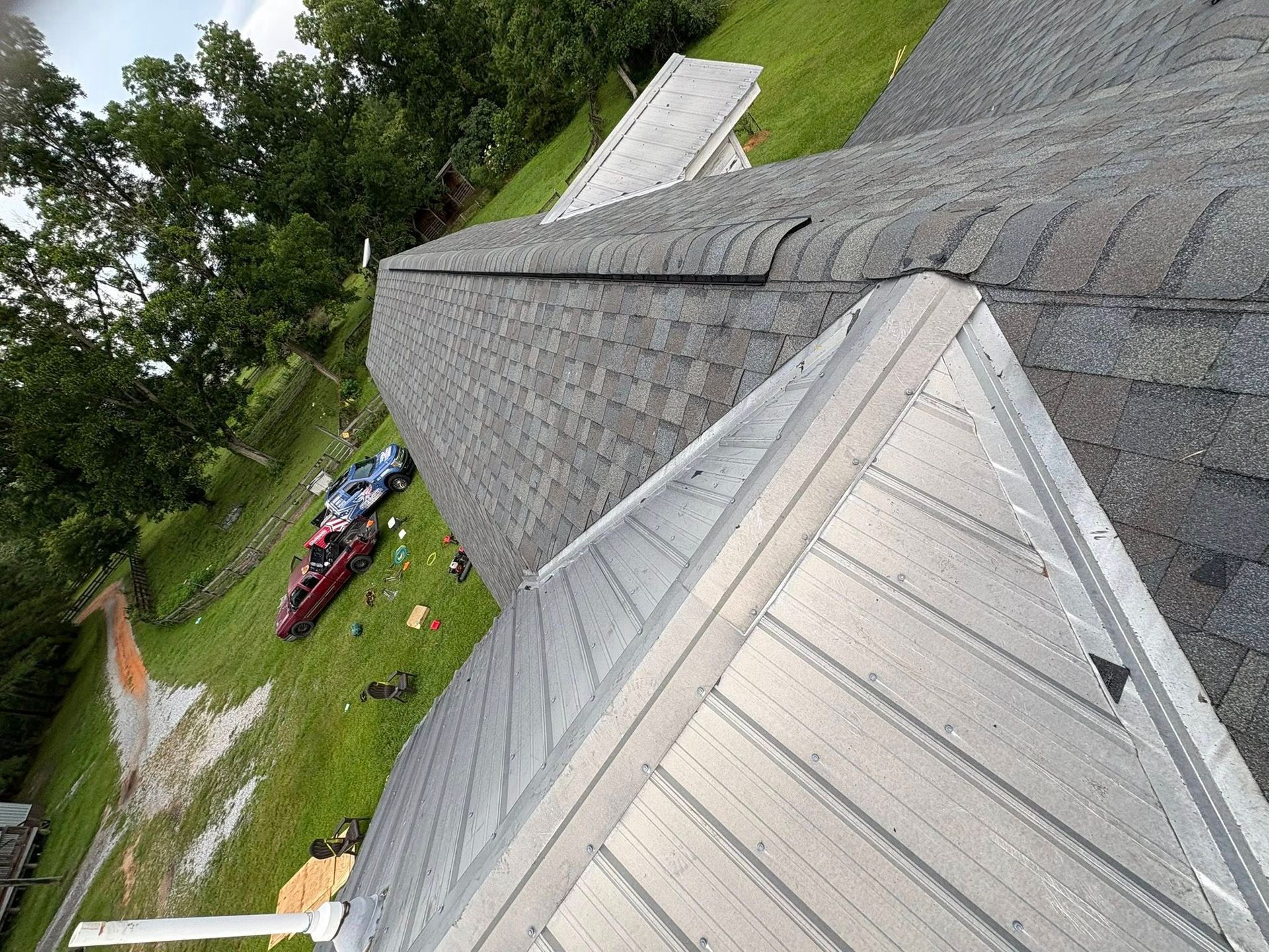 View from above of a roof with grey shingles and metal flashing. Green grass and trees are in the background.