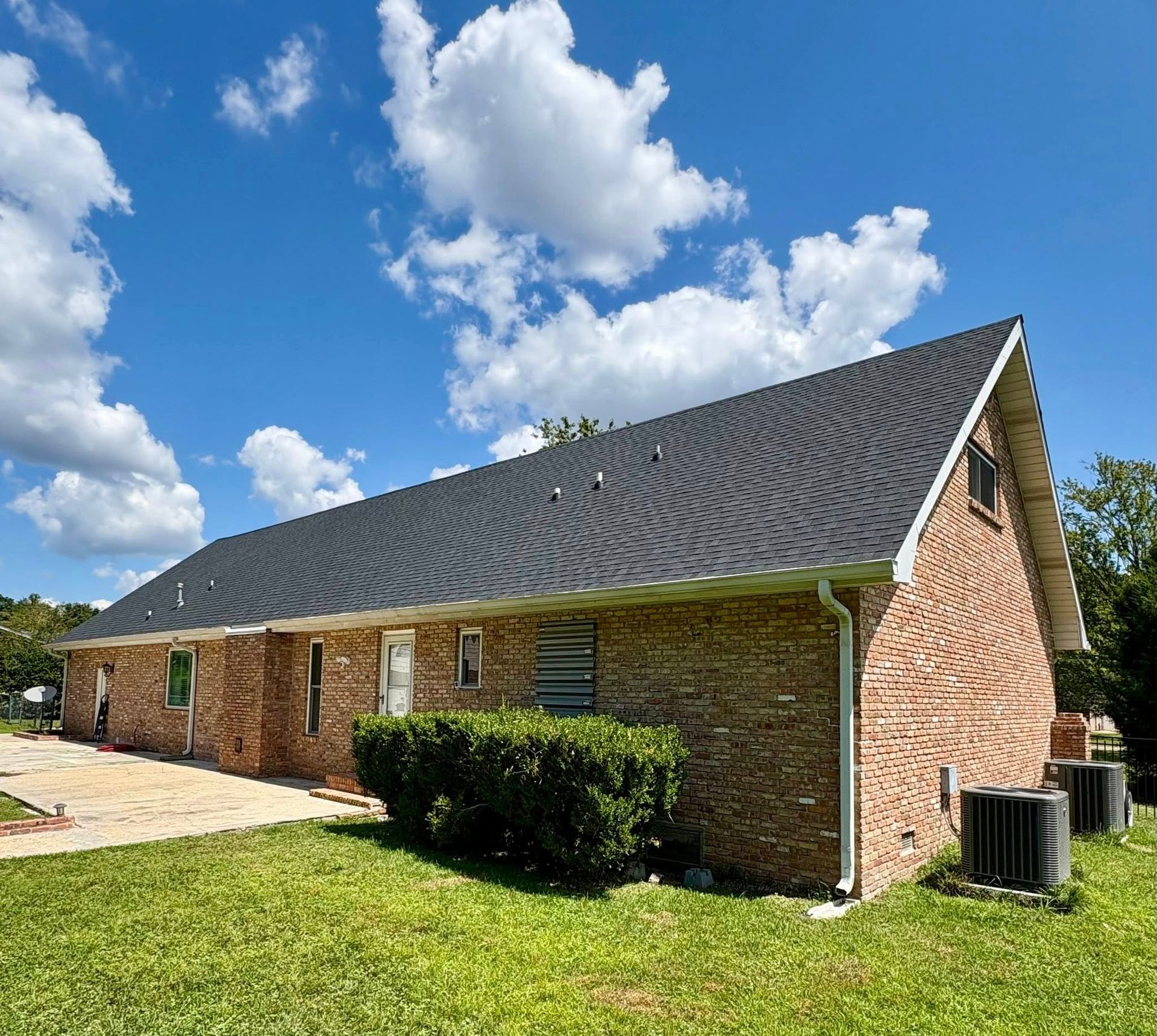 Brick house with black shingle roof, green lawn, and blue sky with clouds.