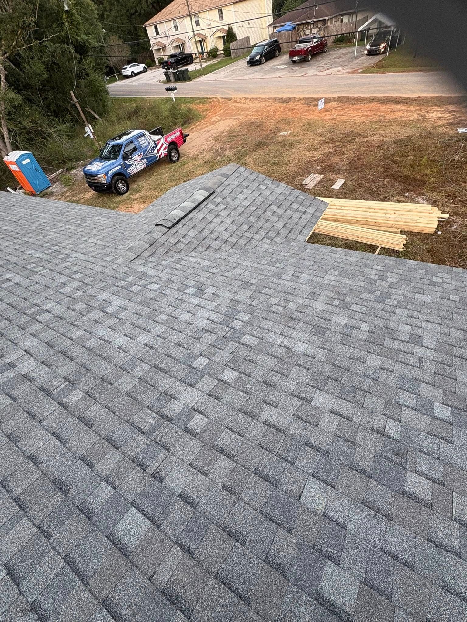 Overhead view of a gray shingle roof under construction, with a truck, porta-potty, and buildings in the background.