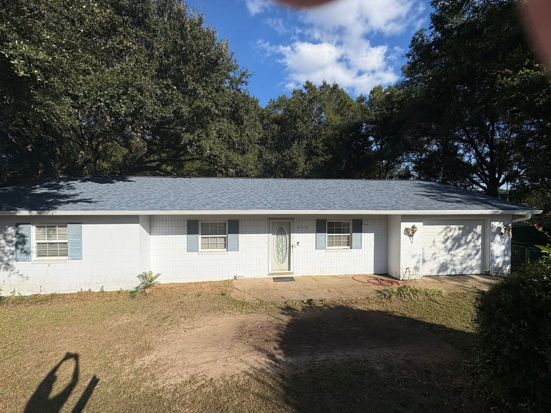 White ranch-style house with blue shutters and roof, set against trees and a cloudy sky.