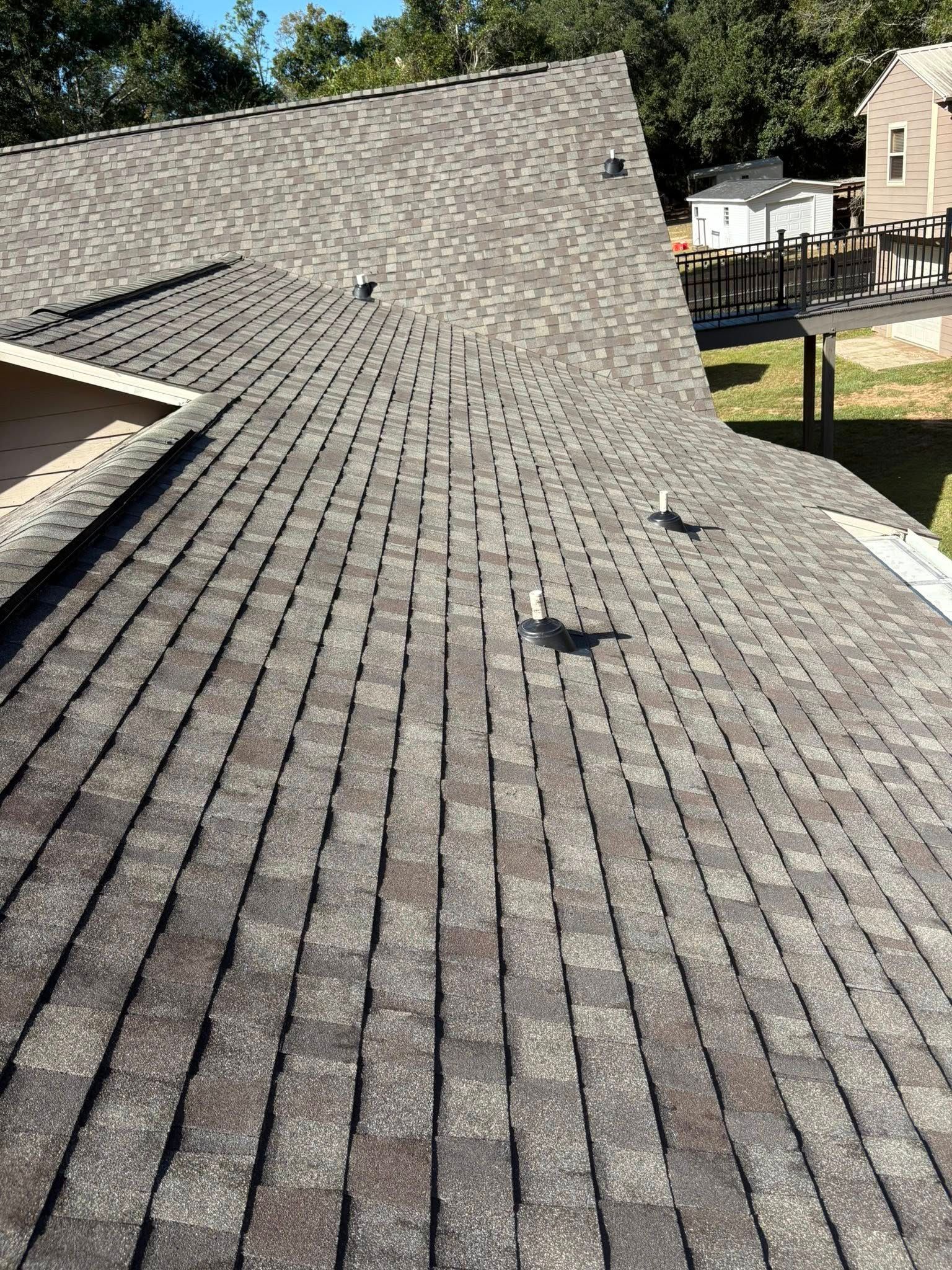 Gray shingle roof on a house, angled view.  Several vent pipes protrude. Sunny outdoor setting.