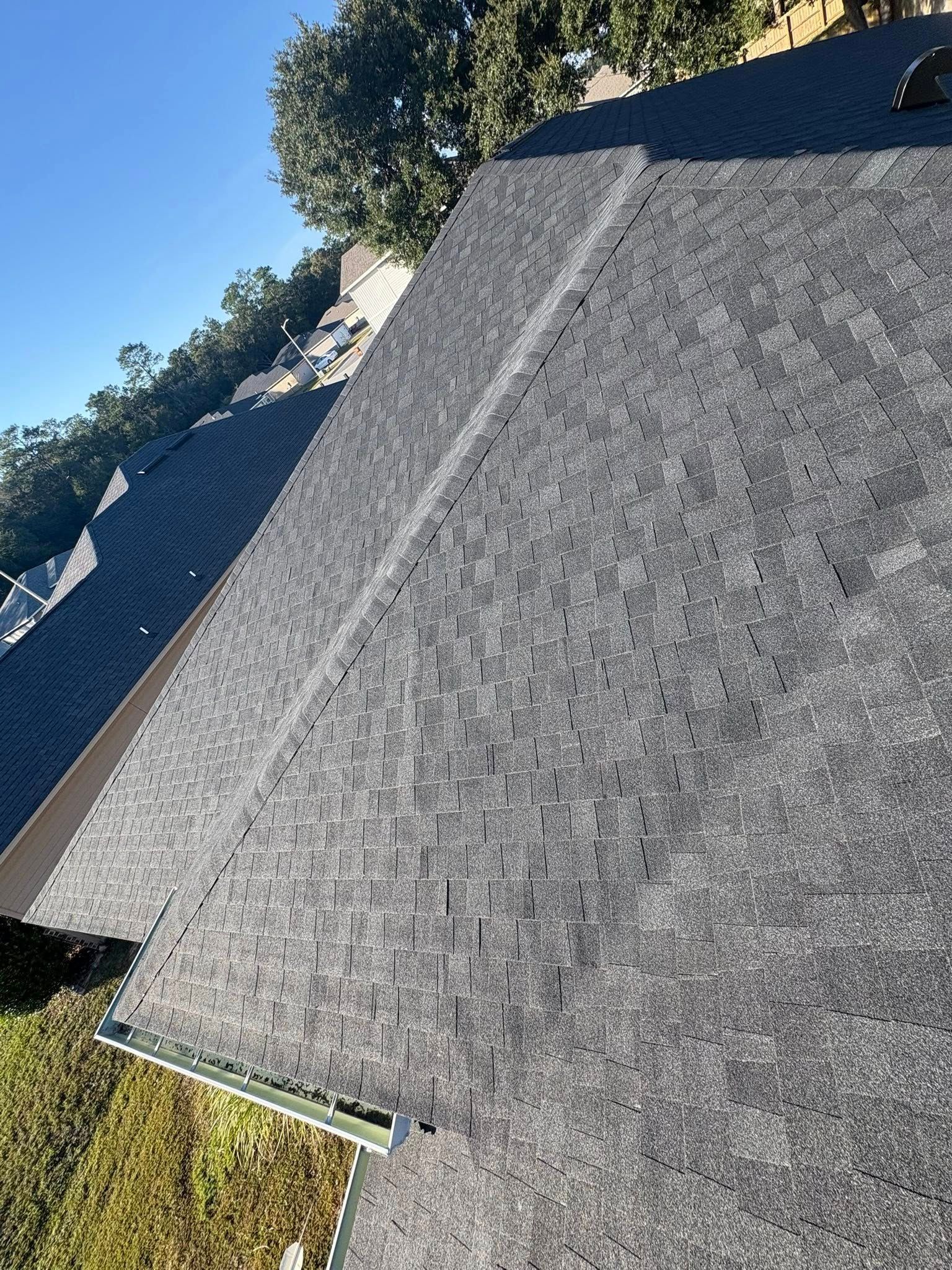 Dark gray shingle roof on a house, angled view from above. Blue sky and green yard visible.