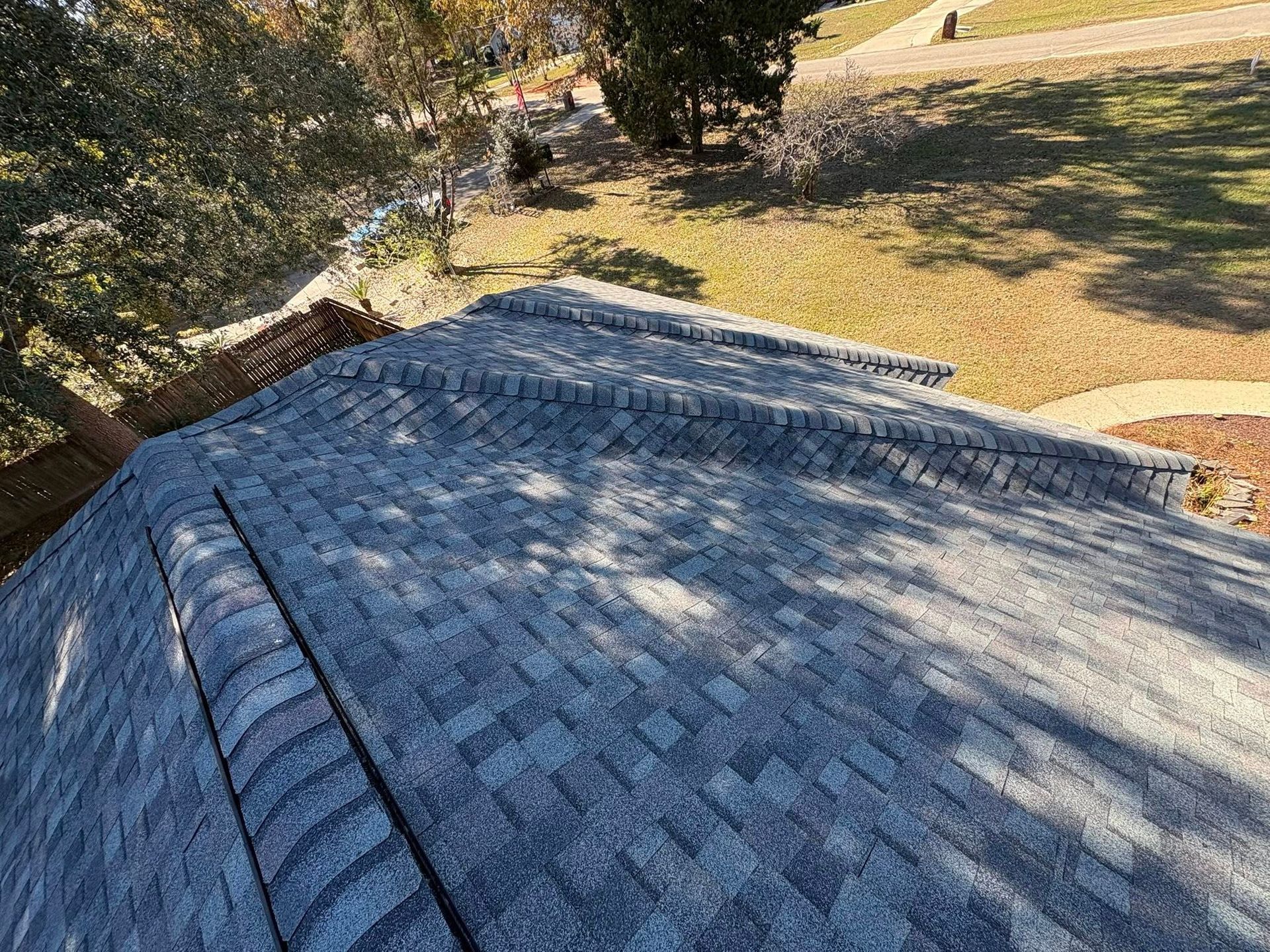 A close-up of a shingled roof with a grassy yard in the background. The shingles are gray and in good condition.