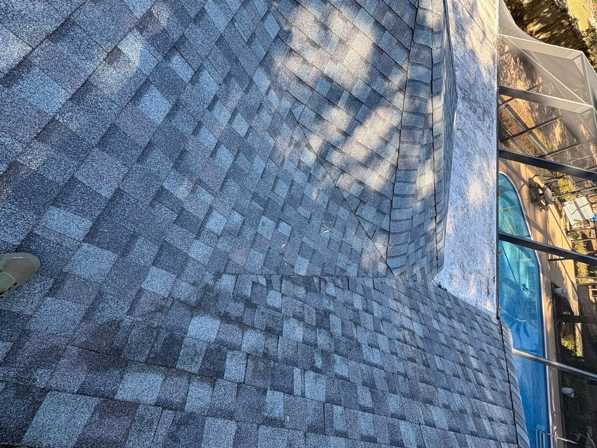 Gray asphalt shingle roof, sunlight casting shadows. A pool and trees are visible to the right.