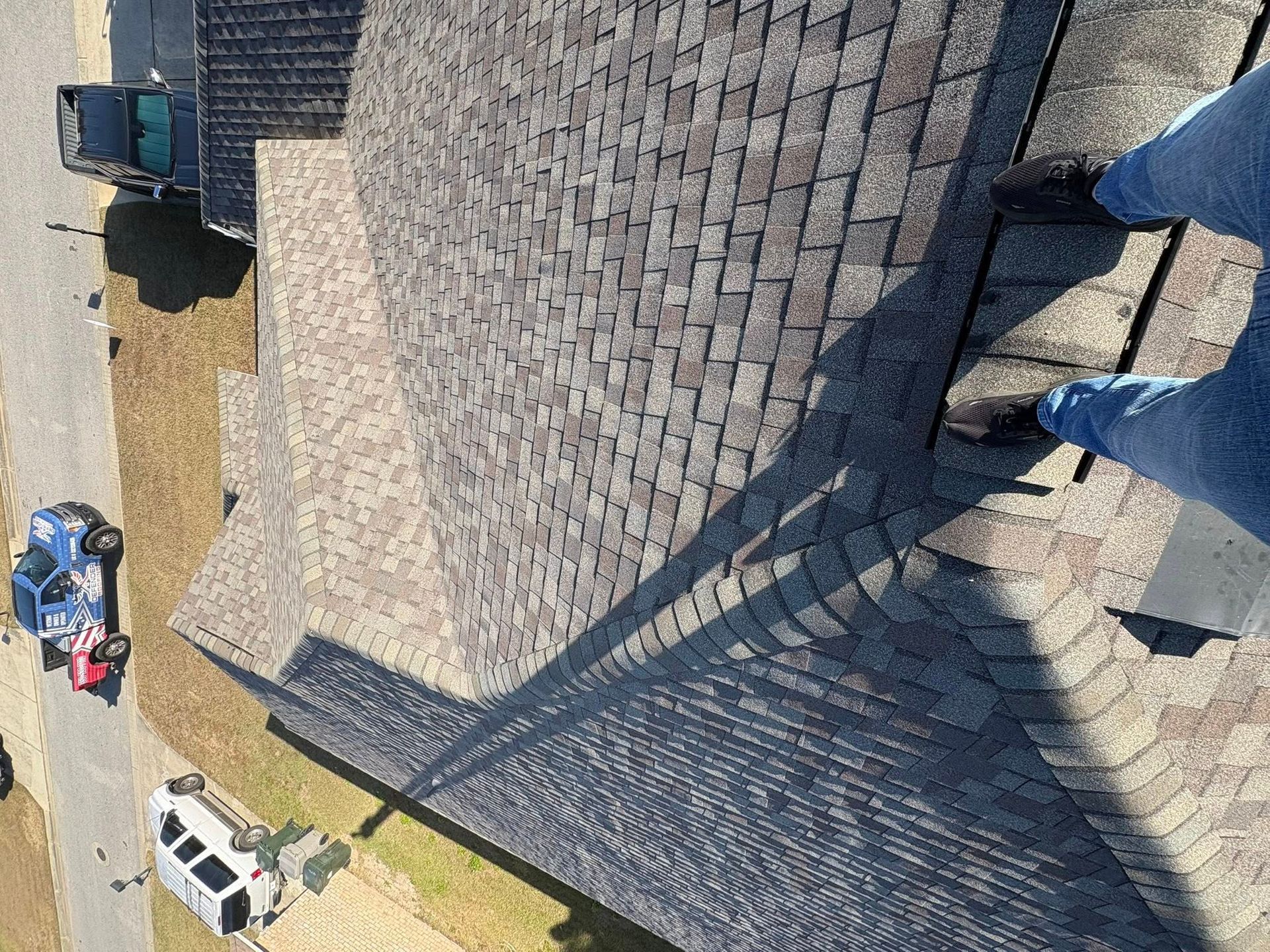 View from above of person standing on a shingled roof, next to a long shadow. Cars are parked below.