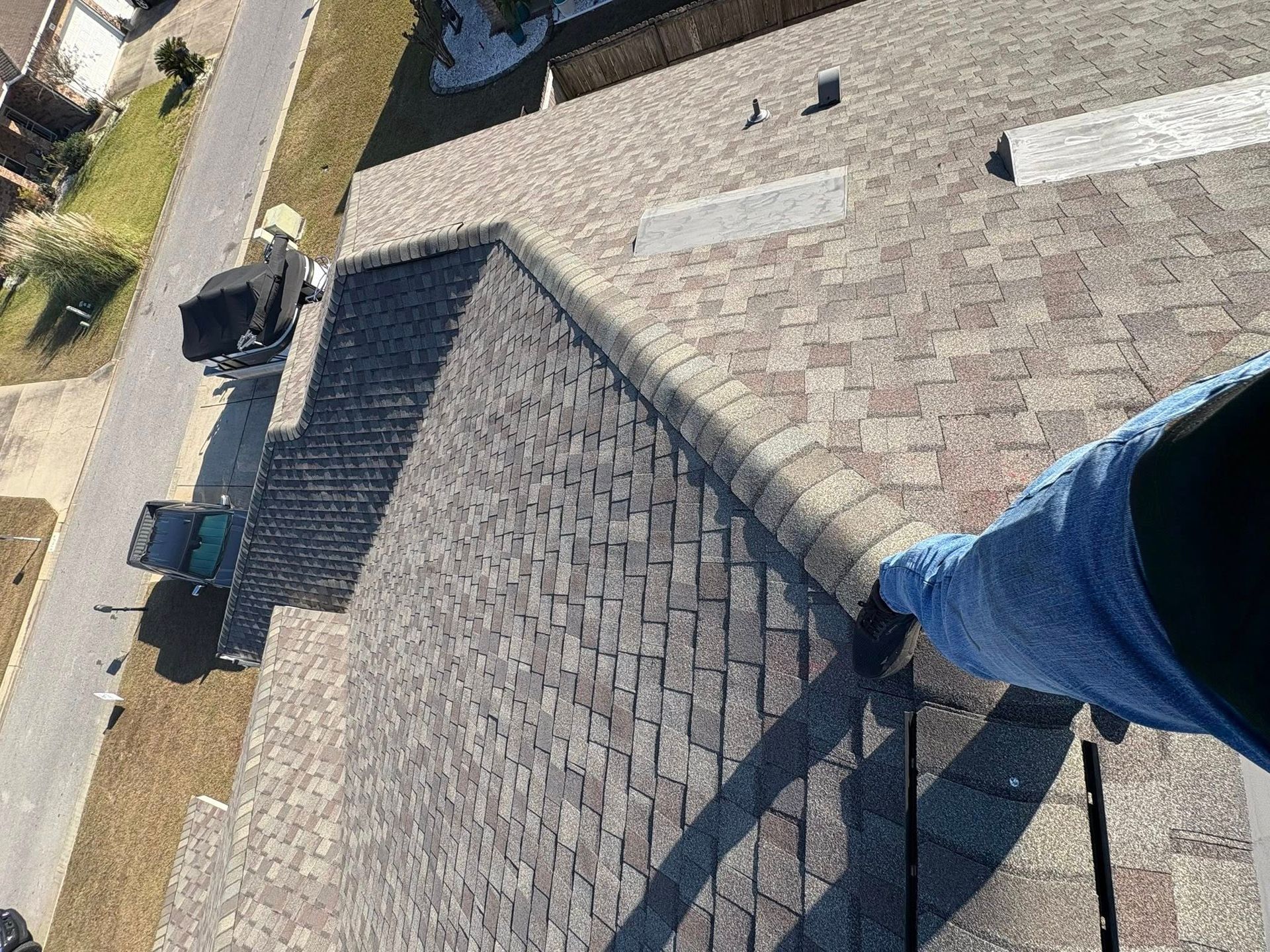 Person on a roof edge, looking down. Asphalt shingles, houses, street, and truck are visible.