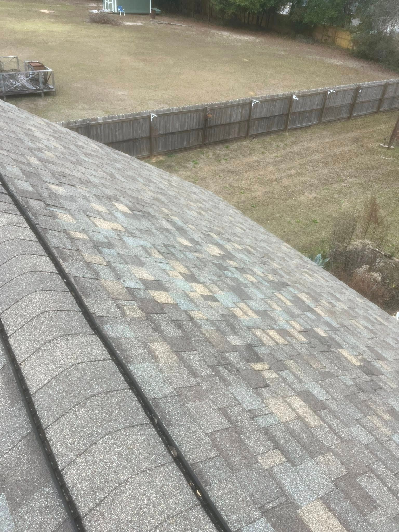 View of an asphalt shingle roof. The roof is weathered with various colors. A wooden fence and lawn are in the background.