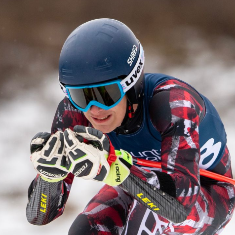 Skier in red and black racing suit, blue helmet and goggles, grips poles, snowy setting.