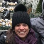 Woman smiling, wearing a black hat with a pom-pom, purple scarf, and black jacket, in a decorated outdoor setting.