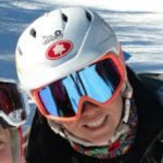 Person in a white ski helmet and orange goggles smiles on a snowy slope.