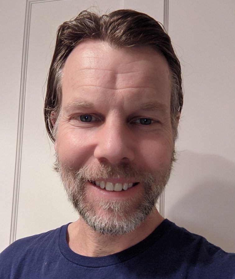 Man with light skin and short brown hair smiles, stubble beard. Wearing a navy blue shirt. White wall behind him.