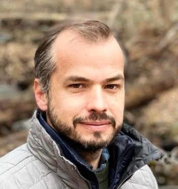 Man with brown hair and beard, wearing a gray jacket, smiling outdoors.