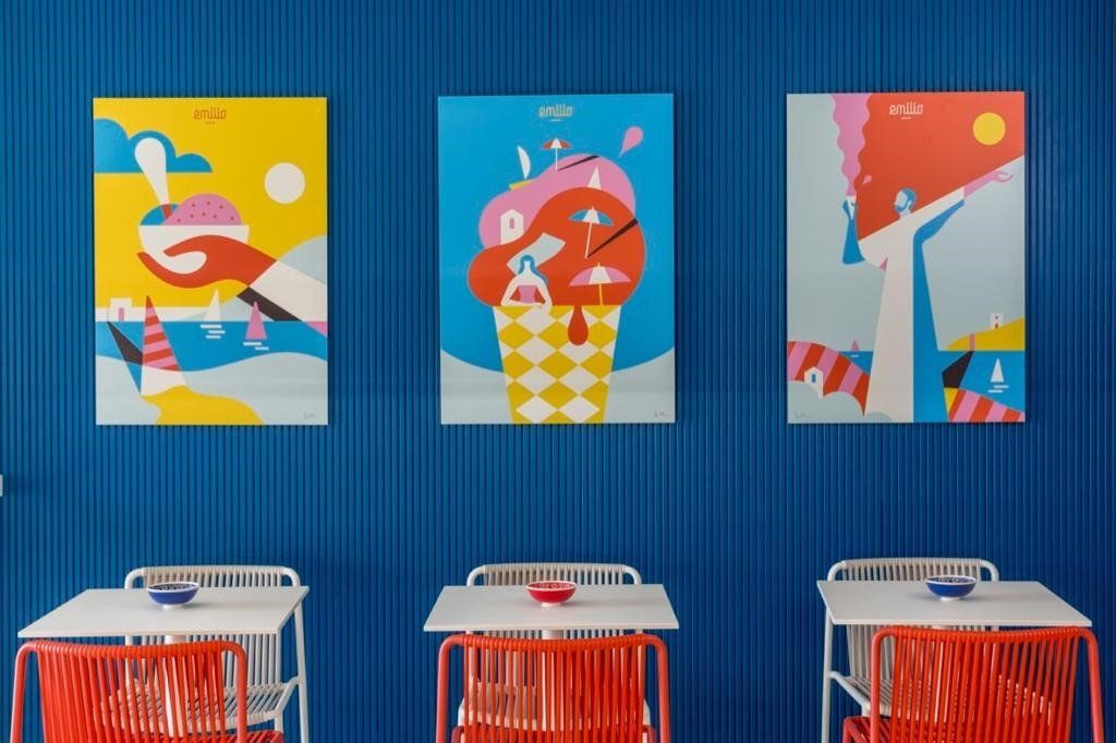 Tre poster colorati di gelati su una parete blu sopra tavoli bianchi con sedie arancioni.