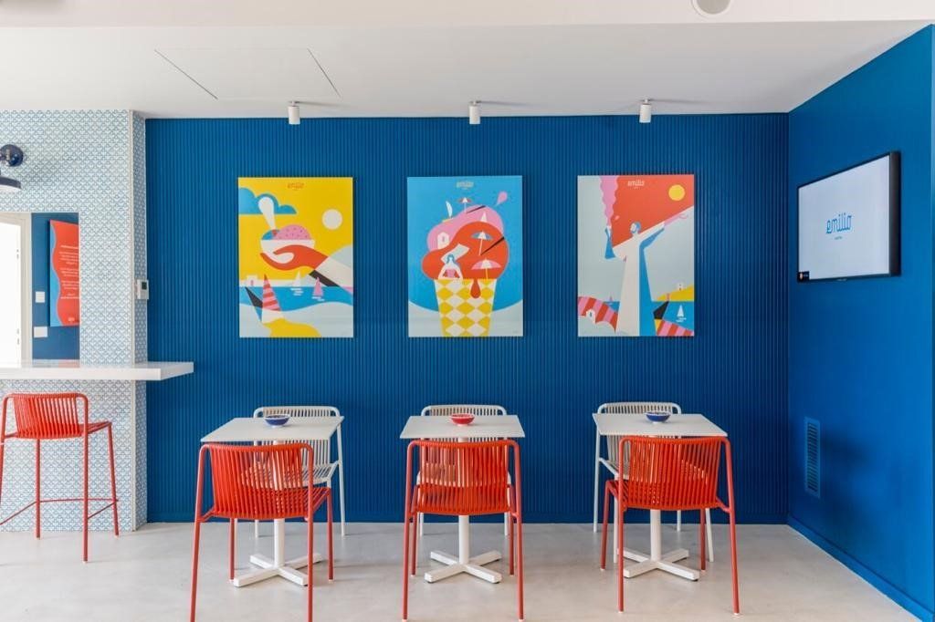 Interno di un negozio con pareti blu, sedie rosse, tavoli bianchi e colorate opere d'arte raffiguranti il gelato.