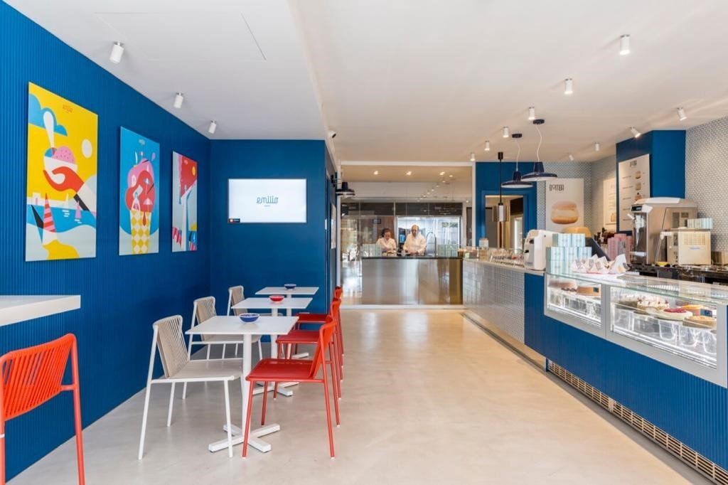 Interno di un bar con pareti blu, tavoli bianchi, sedie rosse e bianche, opere d'arte e un bancone per il servizio.