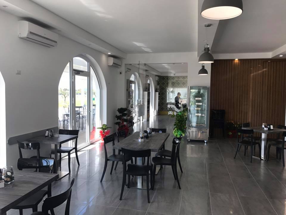Interno di un ristorante moderno con tavoli, sedie e grandi finestre ad arco. Pareti bianche e tavoli grigi.