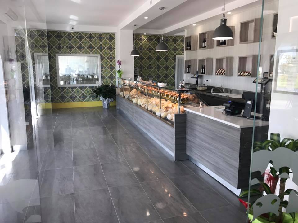 Interno di una panetteria con dolci esposti in una vetrina. Bancone grigio, pavimento piastrellato e parete decorativa.