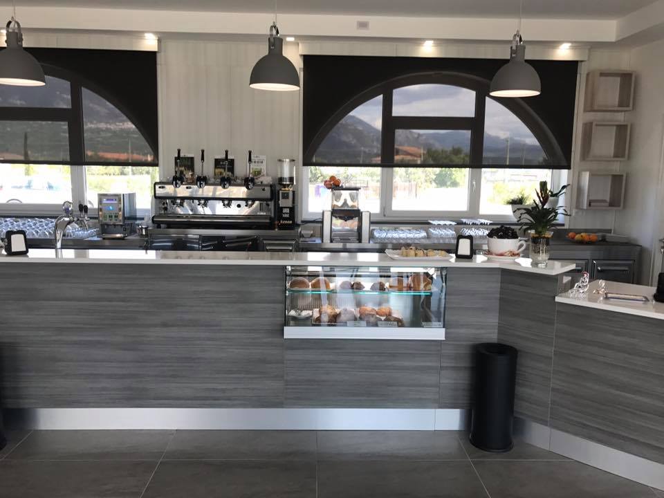 Bancone della caffetteria con macchina per l'espresso, prodotti da forno e finestre ad arco con tende nere.