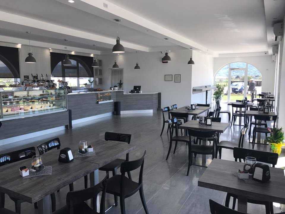 Interno di un bar con tavoli, sedie e una vetrina. Pareti grigie, lampade e una porta d'ingresso in vetro.