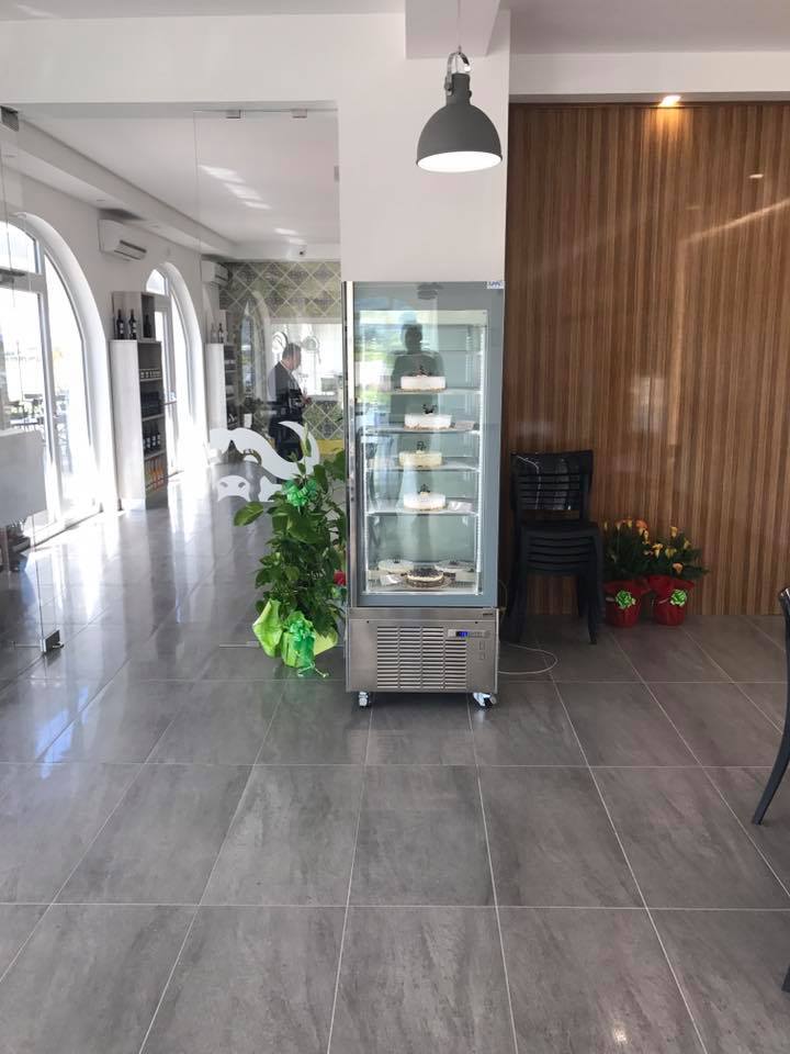 L'interno di un bar con una vetrina refrigerata, una pianta in vaso e una persona sullo sfondo.