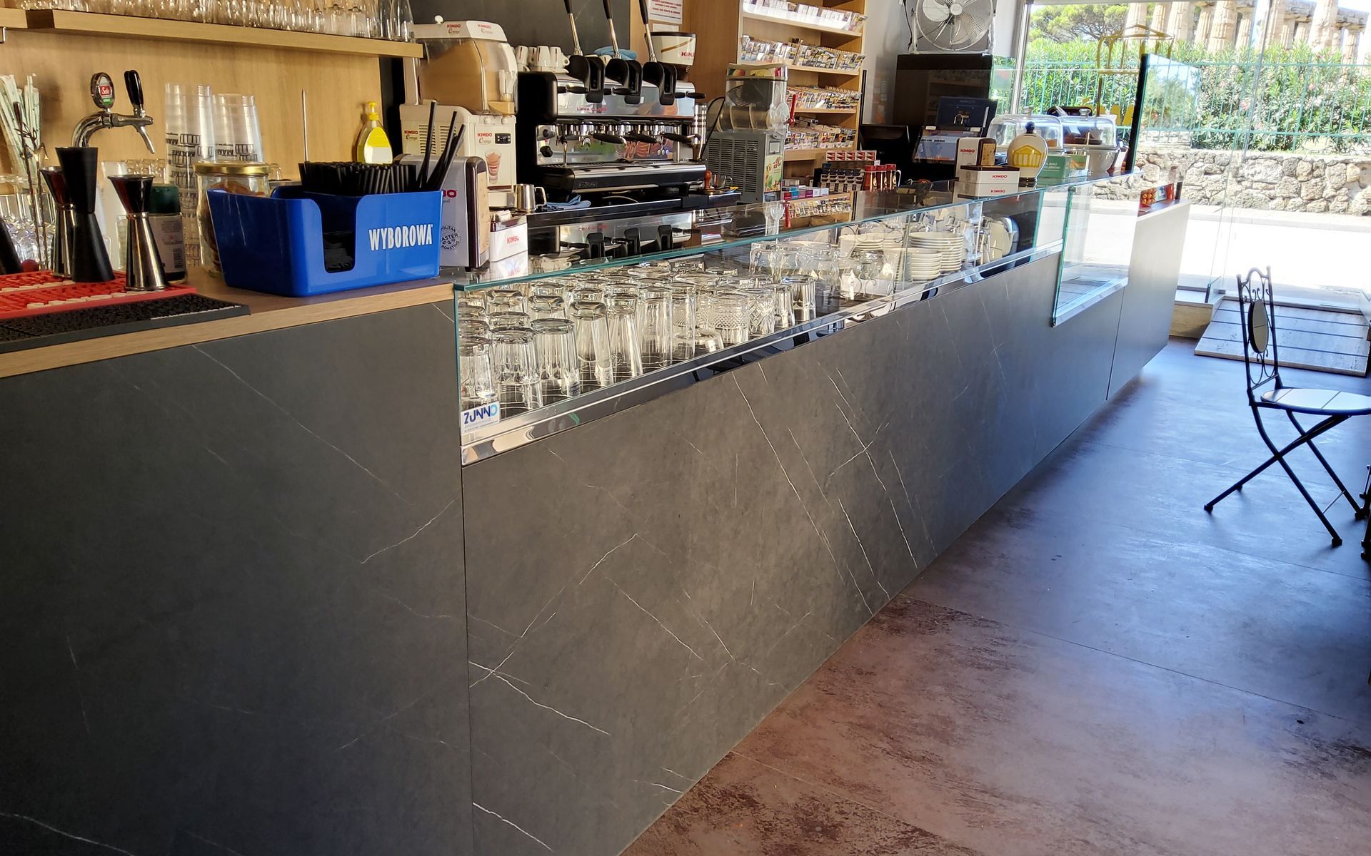 Bancone del bar con bicchieri, bevande e attrezzatura. Bancone grigio con alcune sedie da esterno visibili.